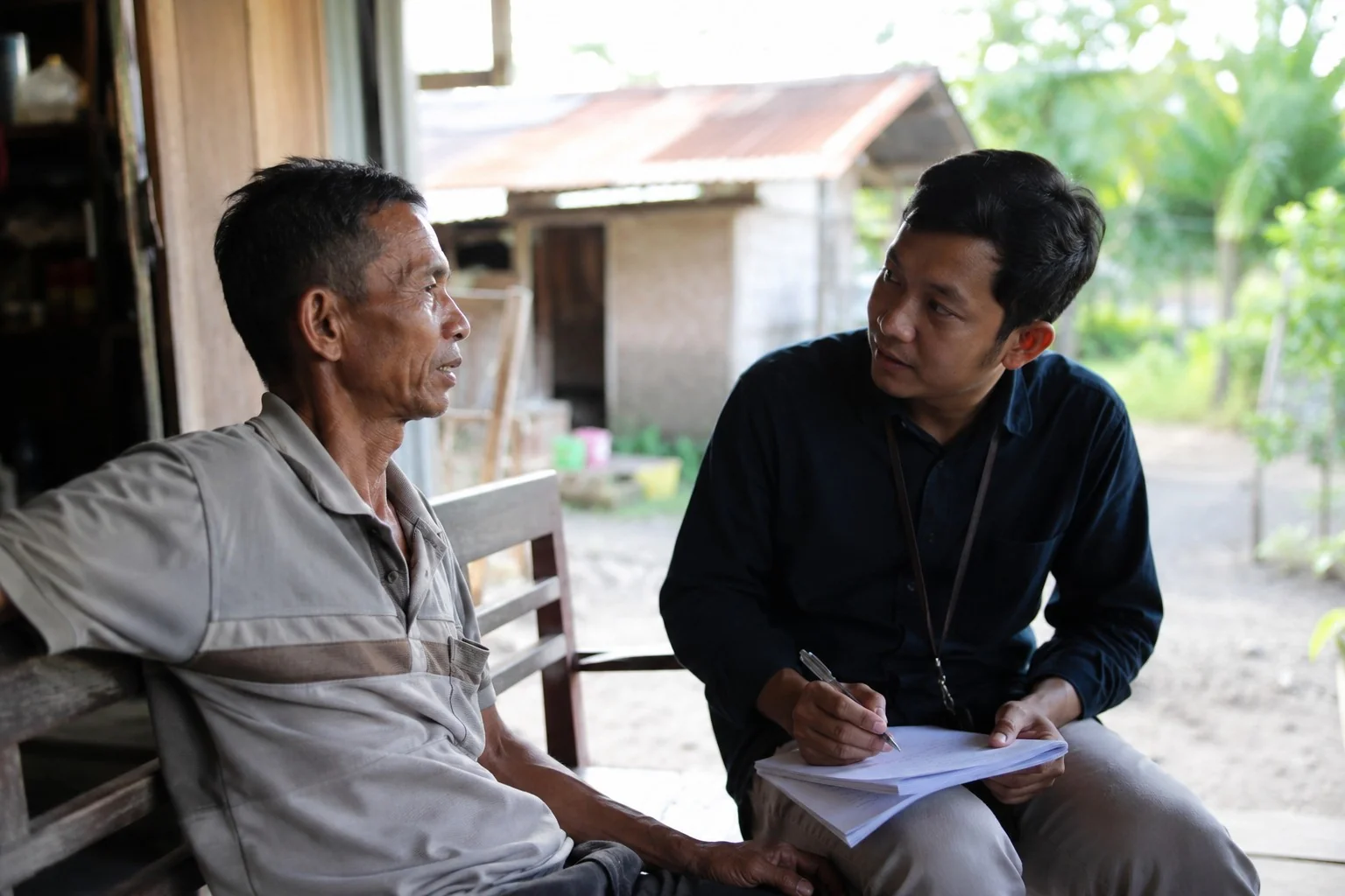 Seorang petugas koperasi muda berbaju hitam mendengarkan dengan saksama keluh seorang anggota lansia di teras rumah kayu di sebuah kampung / A young cooperative officer in a black shirt listens attentively to an elderly member sharing his story on the porch of a wooden village home