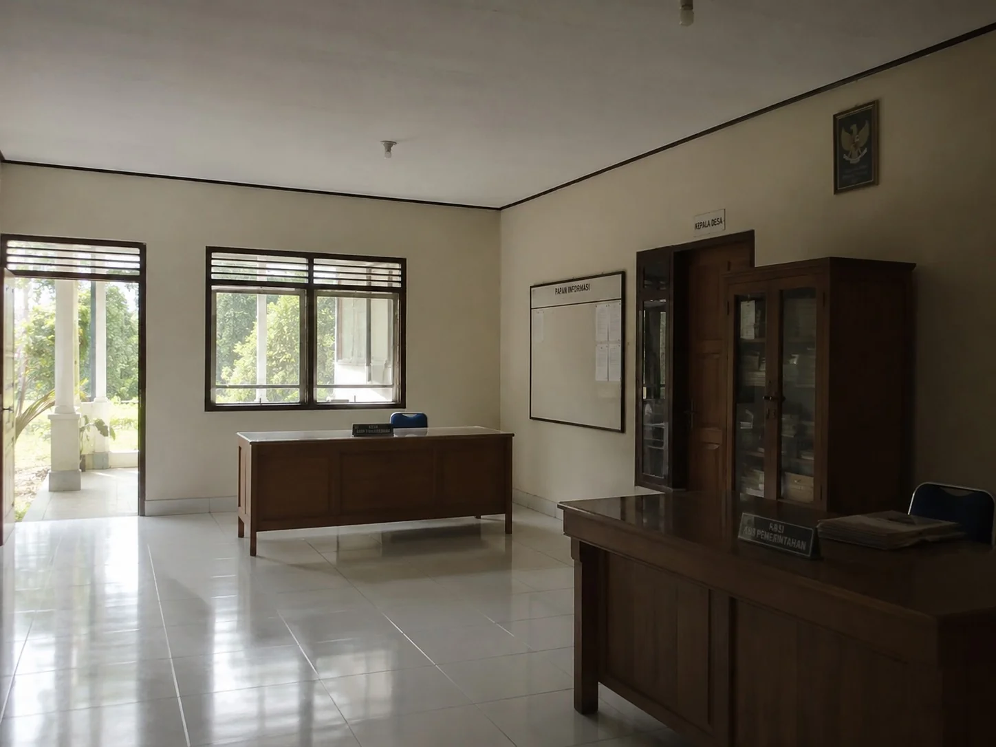Interior kantor desa yang lengang dengan dua meja kerja kayu kosong, papan informasi, dan jendela menghadap pepohonan hijau / Empty village office interior with two vacant wooden desks, an information board, and windows opening to green trees