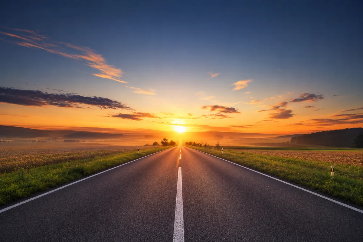 Jalan beraspal lurus membentang menuju matahari terbit di cakrawala di antara hamparan ladang hijau, melambangkan perjalanan menuju masa depan dan harapan yang datang dari Selatan / A straight paved road stretching toward the sunrise on the horizon between green fields, symbolising the journey toward the future and the hope that comes from the South